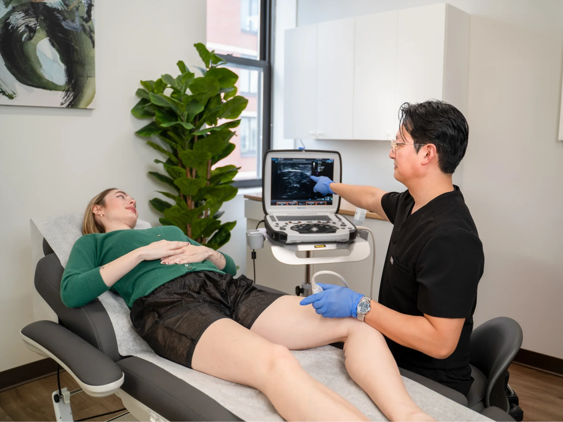 Dr. Benjamin Kum performs a duplex ultrasound on a patient's leg in a clinical setting. He uses an ultrasound probe to examine the area while pointing at the ultrasound machine monitor to explain the diagnostic findings to the patient.