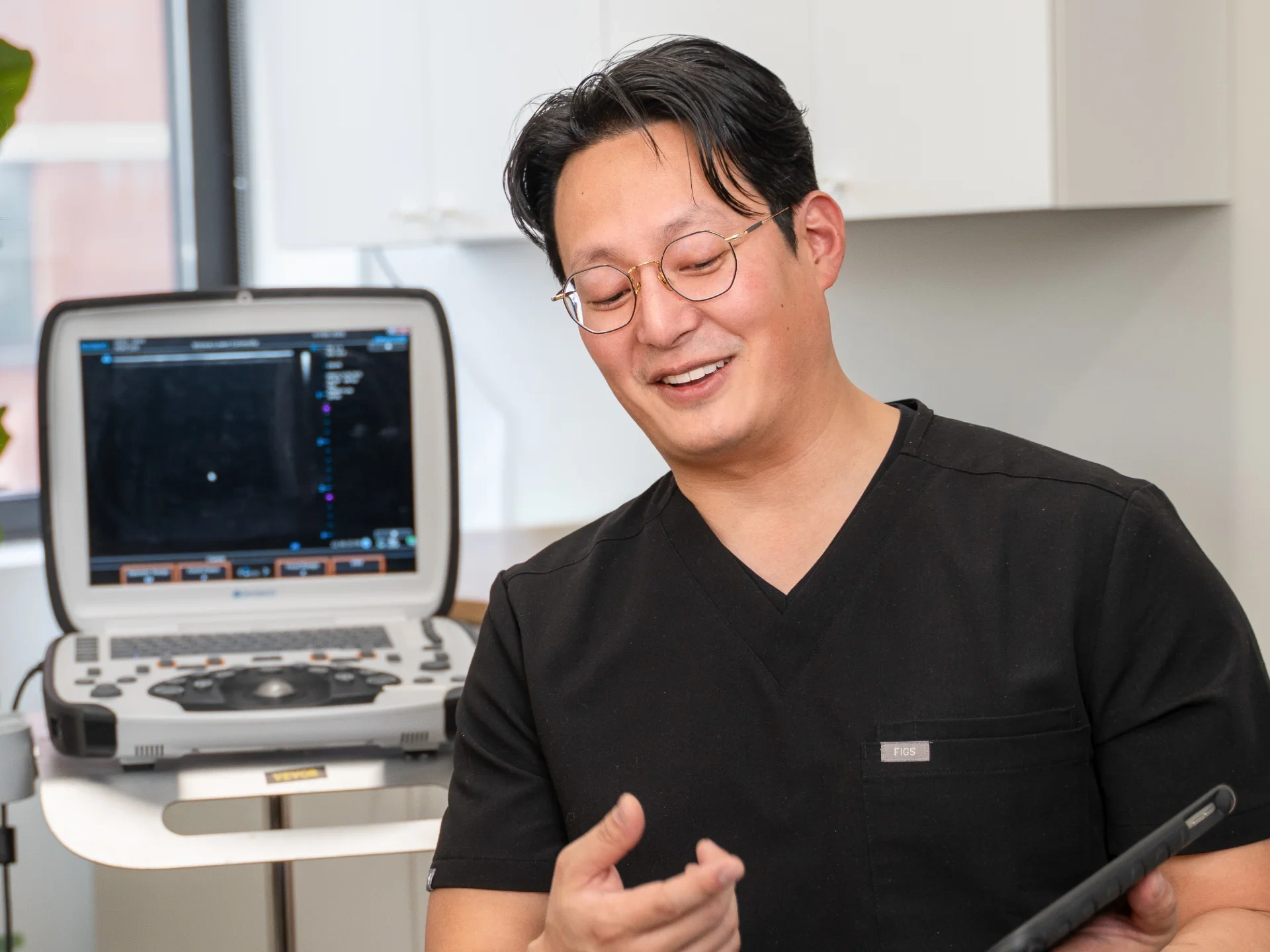 Vein doctor or phlebologist in black scrubs smiling while reviewing information on a tablet, with an ultrasound machine in the background inside a vein clinic.