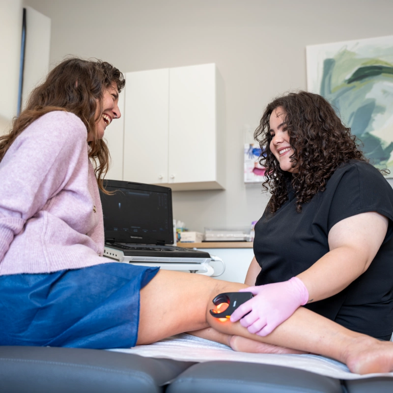 Dr. Hanen Fernandez, MD performs a Duplex Ultrasound examination on a patient's leg using an Ultrasound Machine with an Ultrasound Probe while wearing pink gloves. The patient is seated on an examination table in a clinical treatment room, with ultrasound imaging displayed on a laptop in the background. This diagnostic procedure is used to evaluate venous conditions including Varicose Veins, Spider Veins, Chronic Venous Insufficiency, and Venous Reflux.
